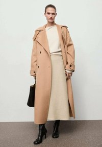 Abrigo de lana color camel con grandes solapas, botones de doble botonadura y correas decorativas en los hombros, combinado con una falda de color crema hasta la rodilla y botas negras hasta el tobillo.