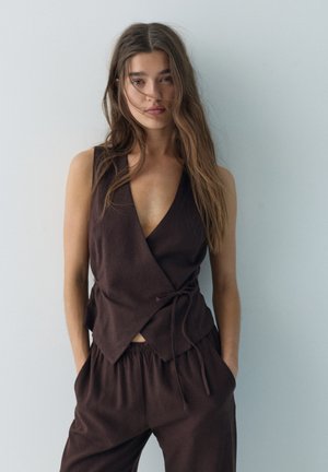 Giovane donna con capelli lunghi indossa un top scuro senza maniche avvolgente e pantaloni larghi abbinati, in piedi contro una parete chiara e semplice con le mani nelle tasche.