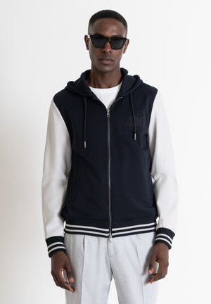 Sudadera con cremallera en color navy y mangas blancas, con cordones y puños a rayas. Logo negro en el pecho y textura suave. Combinada con pantalones ligeros.