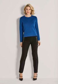 Blonde Frau in einem strukturierten blauen Pullover, schwarzen Hosen mit Reißverschlusstaschen und schwarzen High Heels, die vor einer einfachen hellen Wand steht.