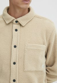 Beige fleece skjortejakke med teksturfinish, med krage, sorte knapper og en brystlomme med et etikett detalj.