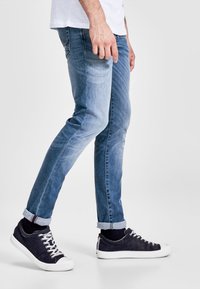 Világoskék farmernadrág, slim fit szabással, enyhe kopással és felhajtott szárakkal. Navy kék cipővel párosítva, fehér talppal és fűzőkkel.