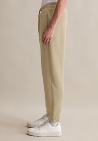 Beige joggingsp pants gemaakt van zachte stof, met een elastische tailleband met een trekkoord, zijzakken en taps toelopende boorden. Draagt witte sneakers.