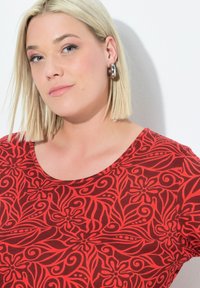 Czerwona bluzka z kwiatowym wzorem i spiralnym wzornictwem. Okrągły dekolt, krótkie rękawy, gładka faktura materiału. Modelka nosi duże srebrne kolczyki w kształcie koła.