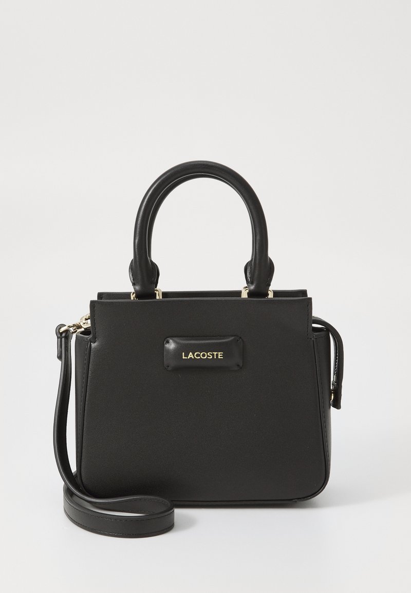 Sac à main Lacoste noir avec un design structuré, texture lisse, poignées supérieures et une bandoulière amovible. Présente un accent de logo doré.