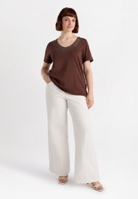 Top marrón de manga corta con escote redondeado y adornos decorativos en los hombros, combinado con pantalones anchos blancos y sandalias con tiras.