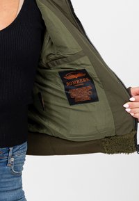 Oliwkowa kurtka bomberka z zamkiem z przodu, miękką polaru podszewką i wewnętrzną metką z nazwą marki "BOMBERS."