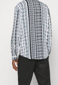 Camicia a maniche lunghe con collo in tessuto nero e bianco, caratterizzata da un motivo di testo ripetuto e onde, con una vestibilità ampia e una leggera lucentezza.
