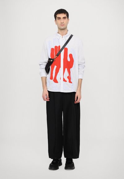 Chemise à carreaux blanche avec un grand motif graphique rouge, associée à un pantalon large noir et des chaussures noires solides. Sac à bandoulière noir porté sur l'épaule.