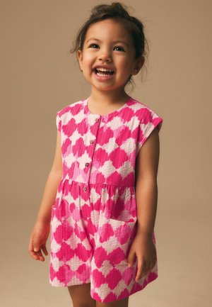 Young girl smiling, wearing a sleeveless pink and white patterned dress with buttons and front pockets, standing against plain beige background.