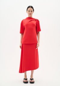 Rotes Oberteil mit kurzen Ärmeln und asymmetrischem Saum, kombiniert mit einem passenden langen Rock aus glattem Stoff, getragen mit schwarzen Sandalen.