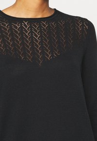 Maglione nero con un motivo in maglia a zigzag trasparente sulla parte superiore del petto e sulle spalle, scollo rotondo, indossato da una persona.