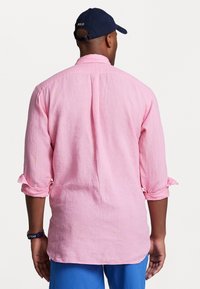 Ljusrosa linnéskjorta med uppvikt ärm, med en veck bak och en avslappnad passform. Tillsammans med blå shorts och en mörk basebollkeps.