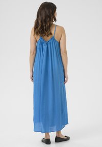 Vestido largo azul con tirantes finos y detalle fruncido en la parte trasera. Tejido ligero, corte suelto y llega hasta los tobillos. Se lleva con zapatos planos negros.