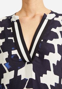 Blůza s výstřihem do V, která má námořnickou modrou a bílou abstraktní vzor, s černobílými pruhovanými detaily na límci. Hladká textura látky.