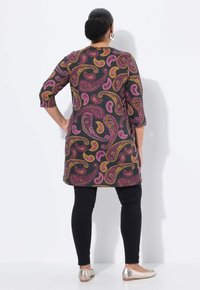 Lange Tunika mit buntem Paisleymuster in Pink und Gelb auf einem dunklen Hintergrund, kombiniert mit schwarzen Leggings und metallischen Ballerinas.