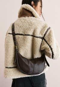 Borsa a spalla in pelle marrone dalla forma curvata, con tracolla regolabile e dettagli in oro, indossata sopra una giacca in pile beige e marrone a trama.