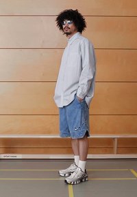 Vaaleansininen raidallinen nappipaita, väljät farkkushortsit, joissa Nike-logo, ja metallinhohtoiset harmaat urheilukengät. Selkeät aurinkolasit täydentävät asua.