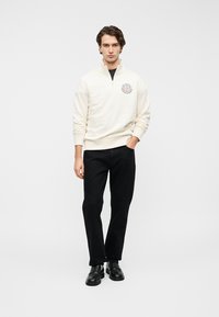 Φωτεινό μπεζ φούτερ με φερμουάρ και στρογγυλό λογότυπο στο στήθος, συνδυασμένο με μαύρο παντελόνι και μαύρα παπούτσια, στέκεται μπροστά σε απλό φόντο.