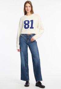 Pull cropped blanc avec le numéro 81 en bleu, associé à un jean large bleu. Tricot texturé et coupe décontractée. Des chaussures noires complètent le look.