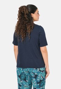 Camiseta de manga corta azul marino con cuello redondo, combinada con pantalones estampados que presentan grandes hojas verdes sobre un fondo oscuro.