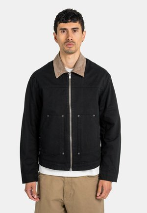Mann trägt eine schwarze Jacke mit Reißverschluss und braunem Kragen, darunter ein weißes Hemd und beigefarbene Hose, steht vor einem einfarbigen Hintergrund.
