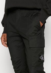Svarta cargobyxor med elastisk midja, sidofickor och ett lapp med Calvin Klein-logotypen på benet. Slätt, strukturerat material.