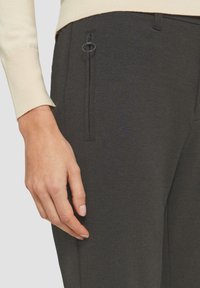 Un pantalon gris foncé à la texture lisse, avec une poche zippée sur le côté dotée d'une tirette circulaire, associé à un pull côtelé de couleur claire.