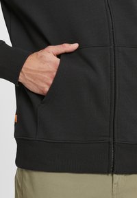 Sudadera con cremallera negra hecha de tela suave, con puños y dobladillo acanalados, bolsillos laterales y una pequeña etiqueta naranja en el lado inferior.