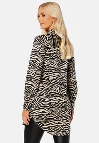 Tunika z długim rękawem w beżowym kolorze w czarne paski zebry, z zaokrąglonym dołem i zapinanymi mankietami, oferująca luźny krój.