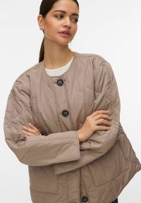 Svetlo bež prešita jakna z velikimi gumbi, okroglim izrezom in dolgimi rokavi. Ima vezen valovit vzorec in stranske žepe.