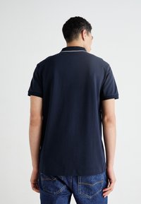 Jūras spēku polo krekls ar īsām piedurknēm, ar klasisku apkakli un kontrastējošu apdari. Kombinēts ar zilām džinsu biksēm.