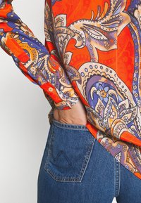 Färgglad mönstrad blus med röda, orange och lila nyanser; lättviktig tyg med synliga knappdetaljer och långa ärmar, kombinerad med denim.