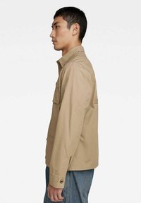 Giacca leggera beige con colletto, due tasche sul petto e dettaglio con bottoni ai polsini. Realizzata in tessuto resistente, vestibilità su misura.