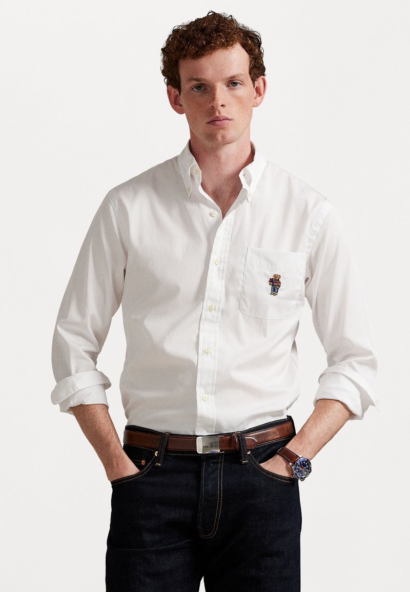 Jauns vīrietis ar kovārtiem sarkaniem matiem, ģērbies baltā krekla ar pogām, uz krūšu kabatas ir mazs izšūts lācis, tumšās džinsās, brūnā jostā un pulkstenī.