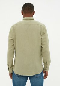 Camisa de botones verde claro con mangas largas, textura suave, dobladillo curvado y puños abotonados, combinada con jeans de mezclilla azul.