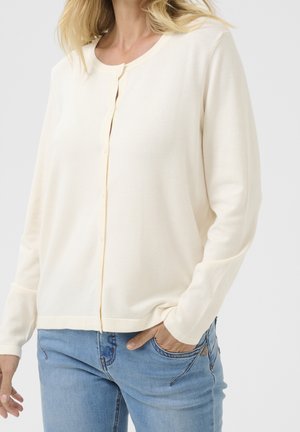 Femme portant un cardigan crème boutonné à manches longues et un jean bleu clair, une main dans la poche sur un fond uni.