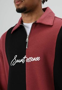 Pull-over bordeaux et noir avec un col zippé, arborant un logo en écriture blanche "Saint essence" sur le devant et une texture en coton lisse.