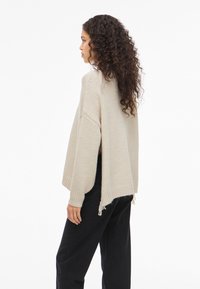 Γυναίκα με μακριά σγουρά μαλλιά, φορώντας έναν χαλαρό μπεζ πλεκτό πουλόβερ και μαύρο παντελόνι, που στέκεται στο προφίλ μπροστά σε έναν απλό λευκό φόντο.