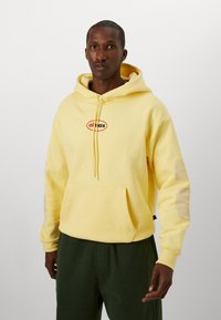 Yellow hooded sweatshirt made of soft fabric, featuring a front pocket and a logo in red and black. Design includes elbow patches and drawstrings.