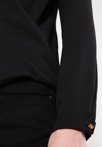 Svart blus med en mjuk, slät textur. Långa ärmar har en manschett med detalj av guldknapp. Enkel, skräddarsydd design.