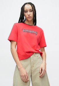 Fiatal személy, fonott hajjal, piros Tommy Jeans pólót és bézs nadrágot viselve, egyszerű fehér háttér előtt áll.