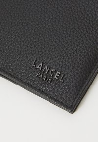 Czarny skórzany portfel o teksturowanej powierzchni, z wytłoczonym logo "LANCEL PARIS". Wyróżnia się kompaktowym kształtem i minimalistycznym designem.