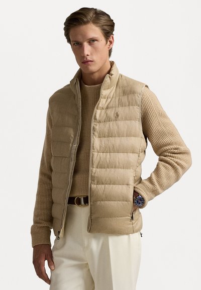 Gilet matelassé beige avec un col haut, fermeture éclair et logo discret. Porté par-dessus un pull texturé crème, associé à un pantalon clair et une montre.