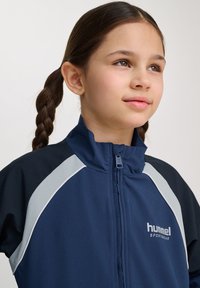 Marinblå sportjacka med hög krage, dragkedjestängning, grå detaljer och textilstruktur. Har en logotyp på bröstområdet.