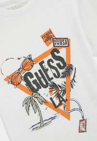 Witte katoenen t-shirt met een grafisch ontwerp van een oranje driehoek, de tekst "GUESS LA", een palmboom, een zonnebril en tags.