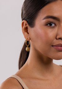 Goldene Creolenohrringe mit einem charmförmigen Sonnendesign, das eine strukturierte Oberfläche aufweist. Das Model hat glattes, dunkles Haar und trägt ein hellfarbiges Oberteil.