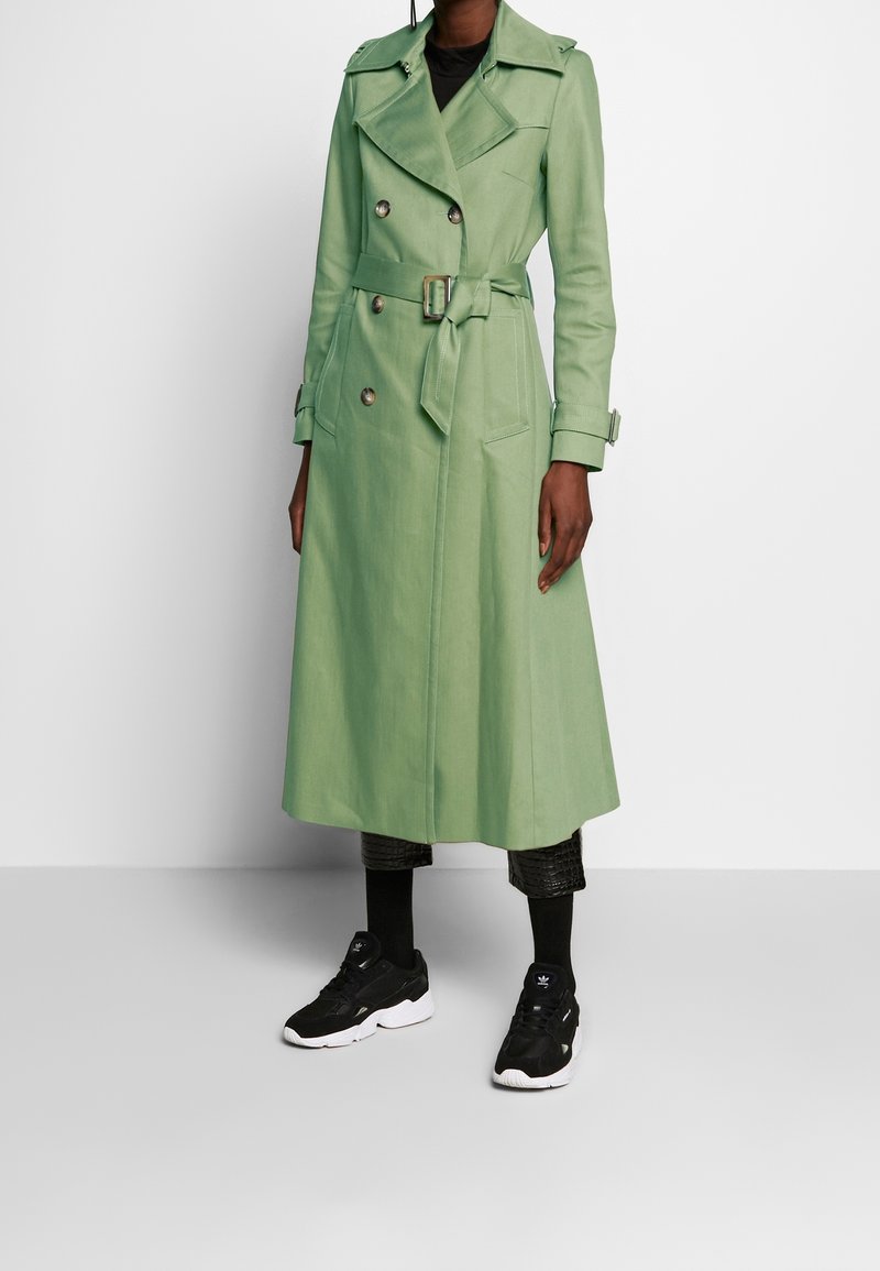 Personne portant un long trench-coat croisé vert clair avec une ceinture, un pantalon noir et des baskets noires à semelles blanches.