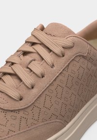 Gros plan sur une basket en daim beige avec des lacets texturés et un motif géométrique perforé sur le côté, semelle beige visible en bas.