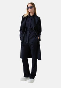 Navy long coat with a belt, layered over a matching shirt and trousers. Features sleek lines and minimalistic design, complemented by white sneakers.
